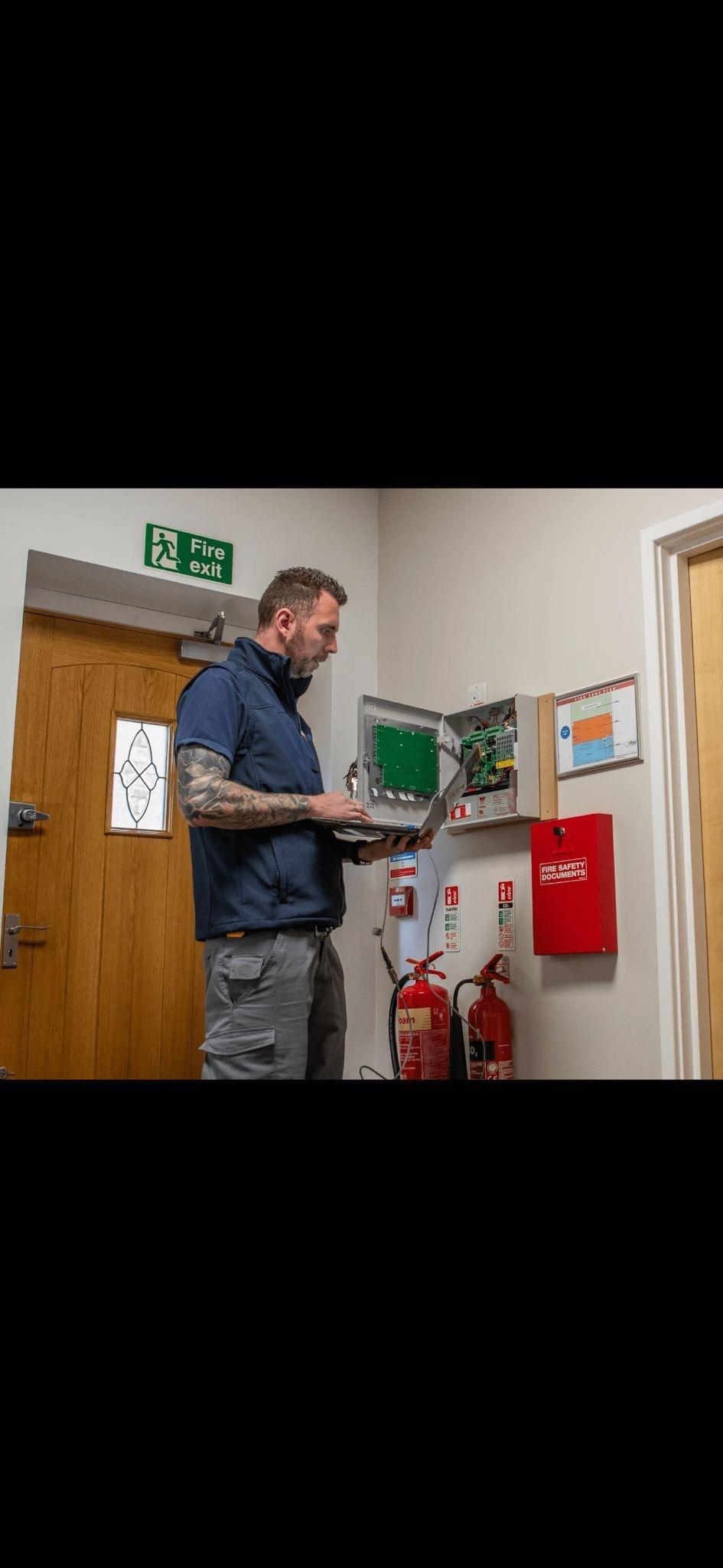 Fire alarm engineer servicing panel with fire extinguishers and exit sign